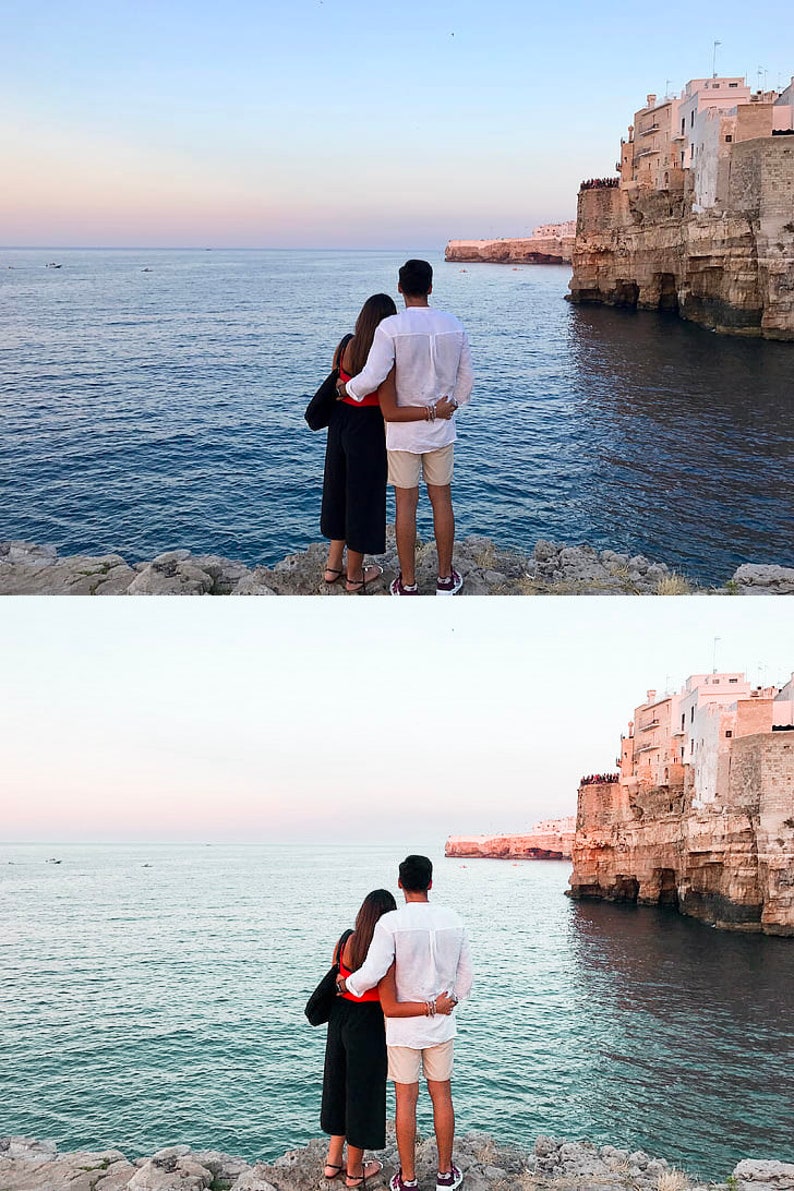 K&ouml;nnte beinhalten: Ein Paar steht auf einer felsigen Klippe mit Blick auf das Meer, im Hintergrund eine in die Klippe gebaute Stadt. Der Himmel ist zartblau mit einem Hauch von Rosa.