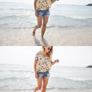 K&ouml;nnte beinhalten: Eine Frau in einem gelben und wei&szlig;en Blusenkleid mit Blumenmuster und Jeans-Shorts geht an einem Sandstrand entlang, im Hintergrund die Wellen des Ozeans.