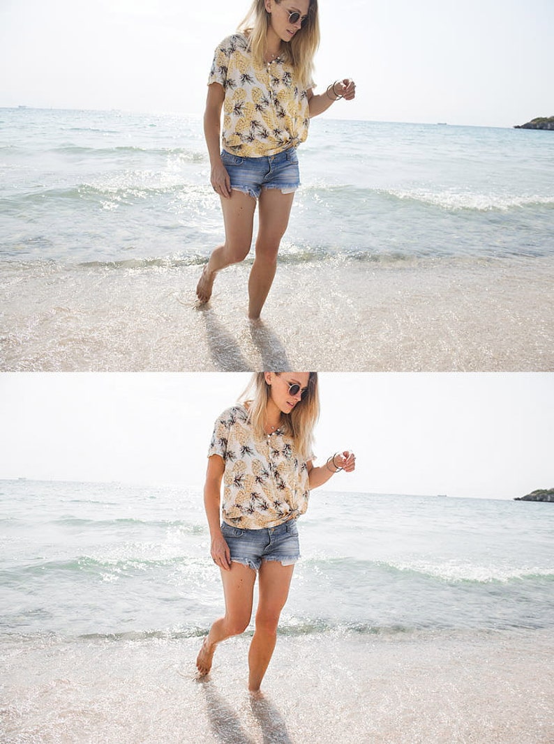 K&ouml;nnte beinhalten: Eine Frau in einem gelben und wei&szlig;en Blusenkleid mit Blumenmuster und Jeans-Shorts geht an einem Sandstrand entlang, im Hintergrund die Wellen des Ozeans.