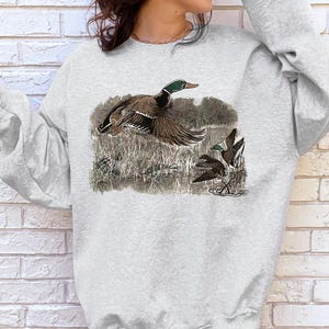 Puede incluir: Una sudadera gris con un estampado gr&aacute;fico de tres patos en un &aacute;rea cubierta de hierba. Los patos son marrones y verdes sobre un fondo blanco.