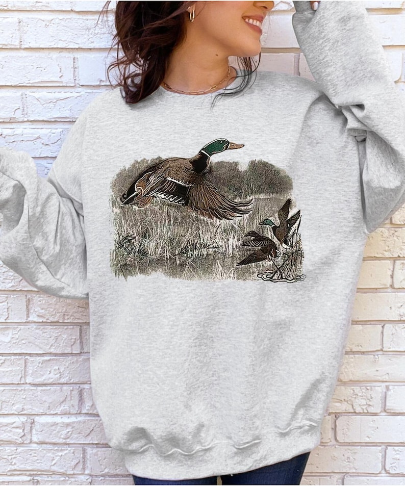 Puede incluir: Una sudadera gris con un estampado gr&aacute;fico de tres patos en un &aacute;rea cubierta de hierba. Los patos son marrones y verdes sobre un fondo blanco.