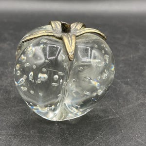 May include: A clear glass paperweight shaped like a peach, with a brass stem and leaf detail. The glass contains numerous air bubbles, creating a unique visual texture. The paperweight is a decorative item.