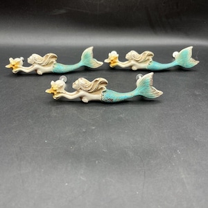 May include: Three drawer pulls shaped like mermaids with cream-colored upper bodies and hair, and teal-colored tails. Each mermaid holds a small yellow starfish in their outstretched hands. The hardware includes silver-toned mounting screws.