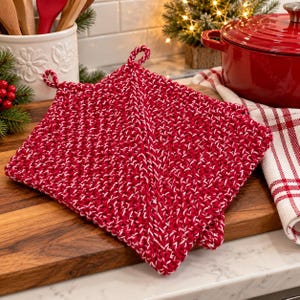 May include: A square, hand-knitted potholder featuring a textured, two-tone pattern of deep red and white yarn. The potholder has a small, integrated loop at the top corner for hanging and a thick, durable construction with a visible diagonal stitch.