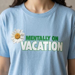 May include: A light blue short-sleeve t-shirt featuring a white daisy flower graphic on the left side and the green and white text "MENTALLY ON VACATION" printed across the chest in a bold, block-style font with a green outline.
