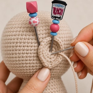 May include: A beige crocheted doll head featuring a circular ear attachment. Two decorative sewing pins are inserted into the head, one topped with a pink cube and blue bead, the other with a coffee cup charm labeled "coffee TEACH repeat".