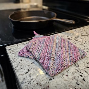 May include: Hand-crocheted potholders in shades of pink, blue, and yellow sit on a kitchen counter next to a cast iron skillet. The potholders are made of yarn and have a textured appearance. Text on the image reads "Kitchen Cozy Hand-Crocheted Potholders."