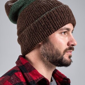 May include: A brown knit beanie with a green stripe near the brim. The hat is made of a thick, textured yarn. The person in the image is wearing a red and black plaid flannel shirt.