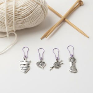 Pode incluir: Um novelo de fio cor de creme, duas agulhas de tricô de madeira e quatro marcadores de pontos com alças roxo-claras. Os berloques prateados incluem um coração com o texto "Crochet Queen", um coração de filigrana, um novelo e uma meada.