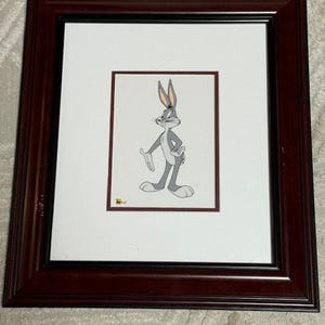 Puede incluir: Obra de arte enmarcada de Bugs Bunny. La imagen presenta a Bugs Bunny en una pose clásica, sobre un fondo blanco. La obra de arte está enmarcada en un marco de madera marrón oscuro con un passe-partout blanco. La obra es de estilo caricatura.