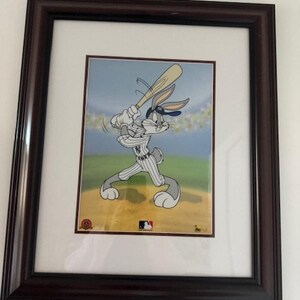 Puede incluir: Obra de arte enmarcada con Bugs Bunny en uniforme de béisbol, bateando. La imagen de dibujos animados está ambientada en un campo de béisbol con un cielo azul. El marco es marrón oscuro y la obra está enmarcada.