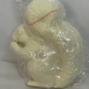 May include: A cream-coloured squirrel figurine, encased in clear plastic. The squirrel is in a seated position, with a textured, rounded body and a fluffy tail. A red line accents the top of the rounded portion. The figurine is likely a decorative item.