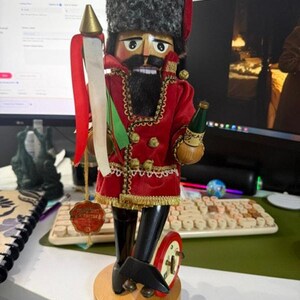 以下が含まれることがあります： 灰色のフェイクファーの帽子、金色のトリムとボタンが付いた赤いベルベットのジャケット、黒いブーツを身に着けた木製のくるみ割り人形。金色の穂先の槍と赤と白のリボン、トカイワインの緑色のボトルを手に持っている。