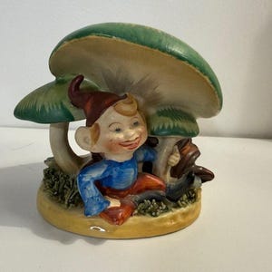 Op de afbeelding: Een keramieken beeldje van een lachende kabouter met een rode hoed en blauw shirt, zittend onder twee groene en beige paddenstoelen. De basis is geel met groen gras.