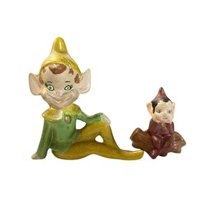 May include: Two ceramic elf figurines. The larger elf wears a yellow hat and green tunic, sitting with legs extended. The smaller elf has a dark brown hat and tunic, sitting on a brown log-shaped base. Both have pointed ears and painted faces.
