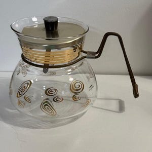 Puede incluir: Una cafetera de cristal transparente con una banda y tapa de metal dorado. La cafetera tiene un asa marrón y está decorada con diseños de remolinos dorados. La tapa tiene un pomo negro. Un artículo de cocina vintage.