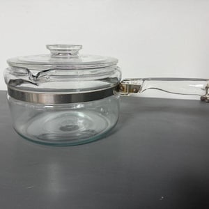 May include: A clear glass saucepan with a matching lid and a clear handle. The saucepan has a silver-colored metal band around the middle. The handle is attached with metal rivets. The lid has a small knob.
