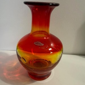 Può includere: Un vaso in vetro soffiato a mano, con un corpo arrotondato e un collo svasato. Il vaso presenta una sfumatura di colori, che passa da un rosso intenso nella parte superiore a un giallo brillante alla base. Il vaso ha un piccolo adesivo trasparente sul davanti.