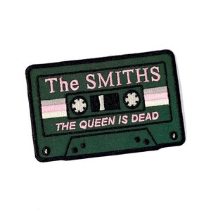 May include: A green embroidered patch in the shape of a cassette tape. The text "The Smiths" is in pink above "THE QUEEN IS DEAD" in pink. The cassette tape design includes a pink, white, and gray stripe.