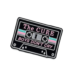 May include: Black embroidered patch in the shape of a cassette tape. The patch features the text "The Cure" in pink and "BOYS DON'T CRY" in white. The cassette tape design includes pink, teal, and gray stripes.
