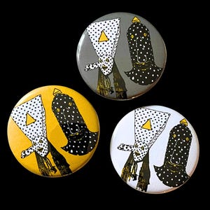 May include: Three round buttons with a black background. Each button features a whimsical design of a figure with a white triangle, black polka dots, and a yellow triangle. The buttons are in yellow, gray, and white.