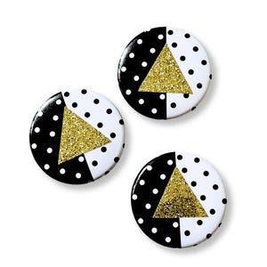 May include: Three round buttons with a black and white polka dot pattern and a gold glitter triangle design. The buttons have a black and white split design with a gold glitter triangle in the center.