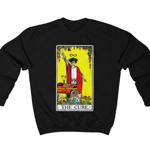 May include: Black sweatshirt featuring a tarot card design with a figure in a red robe, a yellow background, and the text "THE CURE." The design includes a table with a chalice and flowers. The infinity symbol is also present.