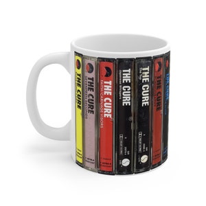 May include: White ceramic mug featuring a design of The Cure album covers. The mug showcases album titles like "Seventeen Seconds" and "Japanese Whispers" in a vertical arrangement. The mug has a white handle and a glossy finish.