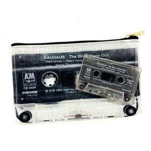 May include: A clear cassette tape and a matching zippered pouch featuring a black and white graphic of a cassette tape. The pouch has the text "BAUHAUS The Sky's Gone Out" and "AM RECORDS" printed on it. The cassette tape is also labeled with the same text.