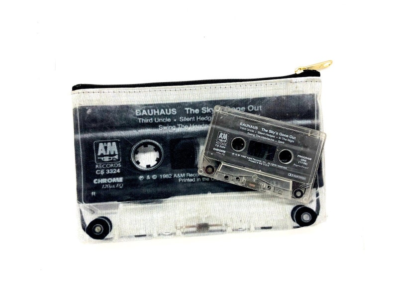May include: A clear cassette tape and a matching zippered pouch featuring a black and white graphic of a cassette tape. The pouch has the text "BAUHAUS The Sky's Gone Out" and "AM RECORDS" printed on it. The cassette tape is also labeled with the same text.
