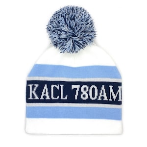 May include: A white and blue knit beanie with a navy blue band featuring the text "KACL 780AM". The hat has a light blue stripe and a navy blue and white pom-pom on top.