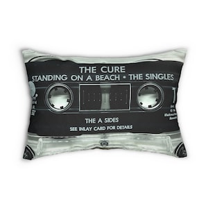 May include: A rectangular pillow featuring a black and white image of a cassette tape. The text on the tape reads "THE CURE STANDING ON A BEACH • THE SINGLES" and "THE A SIDES." The pillow has a clear plastic casing.