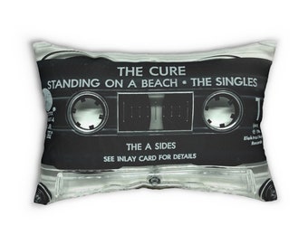 May include: A rectangular pillow featuring a black and white image of a cassette tape. The text on the tape reads "THE CURE STANDING ON A BEACH • THE SINGLES" and "THE A SIDES." The pillow has a clear plastic casing.