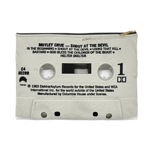 May include: A zippered pouch with a design resembling a cassette tape. The pouch is off-white with black text that includes the band name "Motley Crue" and song titles from the album "Shout at the Devil". A gold-colored zipper is at the top.