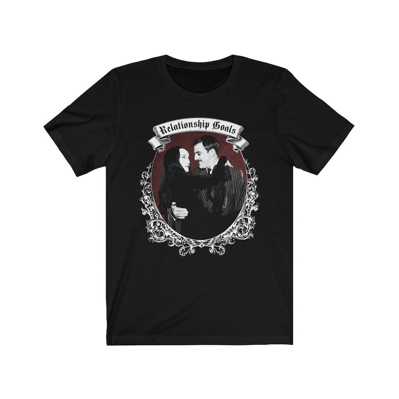 May include: A black short-sleeved t-shirt featuring a graphic print of a couple embracing within an ornate oval frame. Above the couple, a banner displays the text "Relationship Goals" in a gothic font. The design uses black, white, and red.