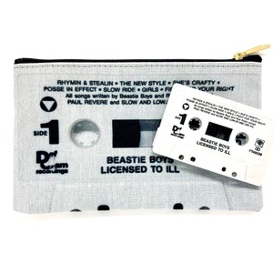 May include: A light gray cassette tape-themed zippered pouch with a gold zipper and a white cassette tape. The pouch features song titles and the text "BEASTIE BOYS LICENSED TO ILL". The cassette tape also has the same text.