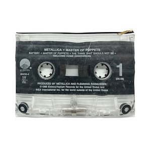 May include: A rectangular zippered pouch featuring a cassette tape design. The pouch has a black and clear plastic appearance with the text "METALLICA MASTER OF PUPPETS" and song titles. The zipper is gold-colored.