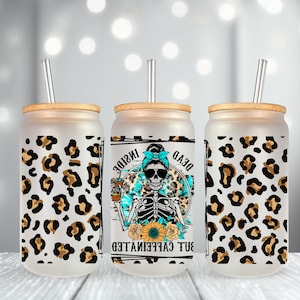 May include: Three frosted glass tumblers with bamboo lids and clear straws. Each tumbler features a different design. The center tumbler has a skeleton wearing sunglasses and a blue bandana with the text "Dead But Caffeinated Inside". The tumblers are decorated with a leopard print pattern.