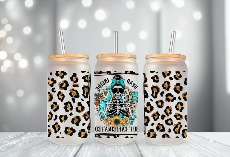 May include: Three frosted glass tumblers with bamboo lids and clear straws. Each tumbler features a different design. The center tumbler has a skeleton wearing sunglasses and a blue bandana with the text "Dead But Caffeinated Inside". The tumblers are decorated with a leopard print pattern.