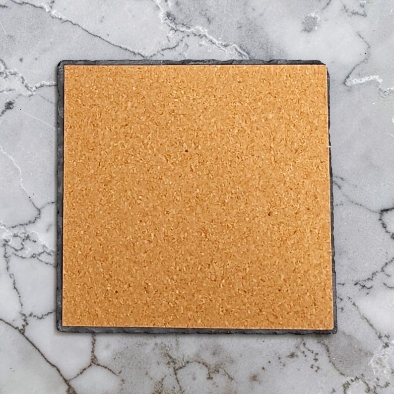 May include: A square coaster featuring a dark gray slate base with a rough, natural edge and a smooth, light brown cork backing centered on the underside. The coaster has a flat, uniform surface and a distinct contrast between the dark stone and cork.