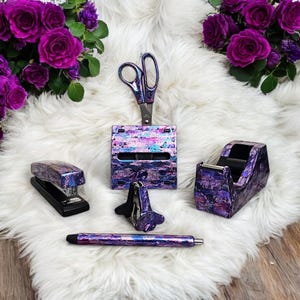 May include: A collection of office supplies, including a stapler, scissors, tape dispenser, pen, and a paper clip holder. The items have a purple, blue, and silver iridescent finish. The items are arranged on a white, fluffy surface, with purple roses in the background.