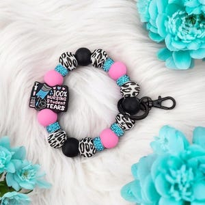May include: A pink, black, and blue beaded keychain with a charm that says "100% Nursing Student Tears".