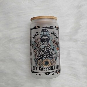 May include: A clear glass jar with a bamboo lid and a black and white graphic of a skeleton wearing sunglasses and a blue bandana. The text on the jar reads "Dead Inside But Caffeinated".