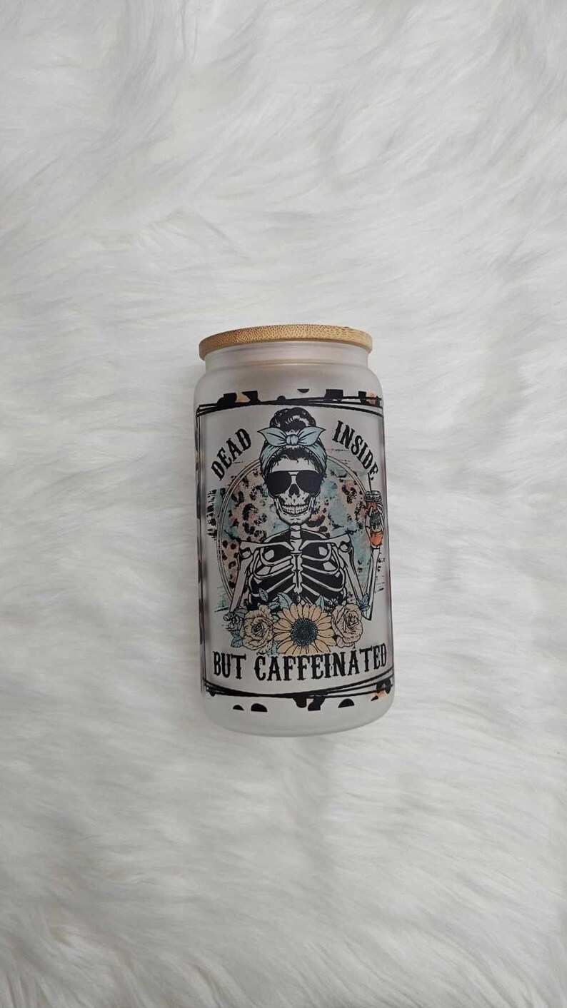 May include: A clear glass jar with a bamboo lid and a black and white graphic of a skeleton wearing sunglasses and a blue bandana. The text on the jar reads "Dead Inside But Caffeinated".