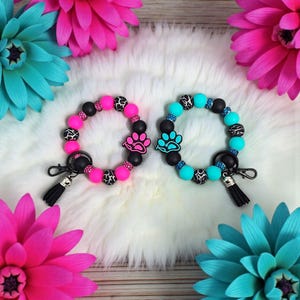 May include: Two beaded keychains with paw print charms. One keychain is pink and black, the other is teal and black. Both have a black tassel and a silver clasp. The keychains are surrounded by pink and teal flowers.