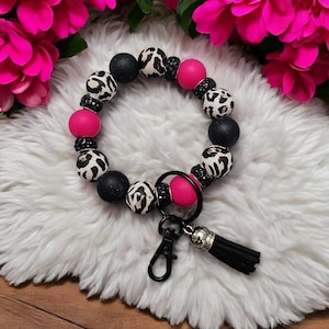 May include: A black and white leopard print beaded keychain with pink beads and a black tassel. The keychain has a silver ring and a black metal clasp.