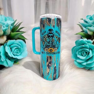 May include: A turquoise and black stainless steel tumbler with a handle. The tumbler has a graphic of a skeleton with a coffee cup and the text "Dead Inside But Caffeinated".