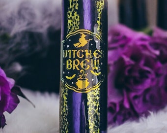 May include: A tall, cylindrical tumbler with a black and yellow pattern and the words "Witches Brew" in gold lettering. The tumbler features a witch on a broom and a cauldron graphic. The background includes purple flowers and candles.