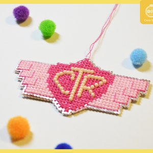 Puede incluir: Un diseño de punto de cruz rosa y blanco con las letras "CTR" en el centro. El diseño tiene forma de corazón con alas. El punto de cruz está colgado de un hilo blanco.