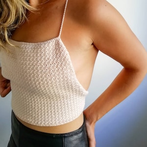 Puede incluir: Un top corto de crochet blanco con tirantes finos. El top es de corte holgado y tiene un escote cuadrado. La modelo lleva una falda de cuero negra.